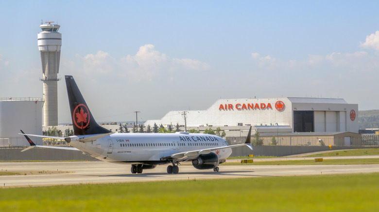 Ein Boeing 737 MAX 8-Flugzeug von Air Canada rollt auf der Landebahn, im Hintergrund ein Flugsicherungsturm und ein Air Canada-Hangar.