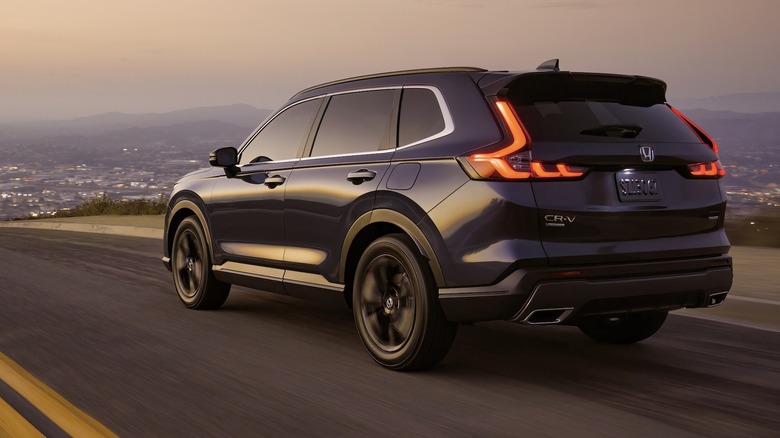 3/4-Hinteransicht eines schwarzen 2024 Honda CR-V, der in der Abenddämmerung eine Straße entlang fährt, mit einer Stadt und Bergen im Hintergrund