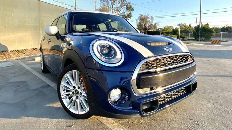 Eine Dreiviertelaufnahme von vorne von meinem blauen Mini mit weißen Streifen, der auf einem Parkplatz vor einem Maschendrahtzaun und einem blauen Himmel geparkt ist