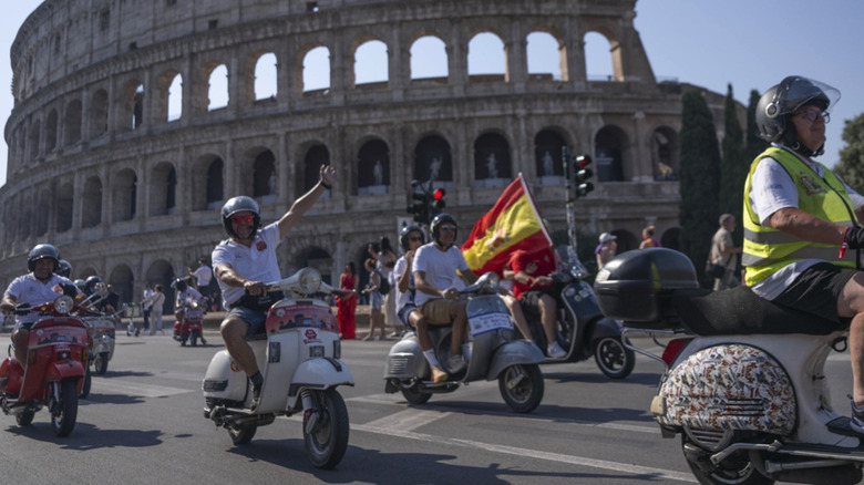 Ein Mann mit Helm auf seiner weißen Vespa winkt der Kamera zu, umgeben von anderen Vespa-Fahrern, die durch das Kolosseum in Rom fahren.