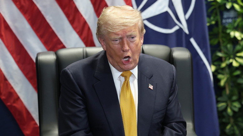 Präsident Trump sitzt möglicherweise mitten im Satz auf einer Konferenz auf einem Stuhl und hat die amerikanische Flagge hinter sich