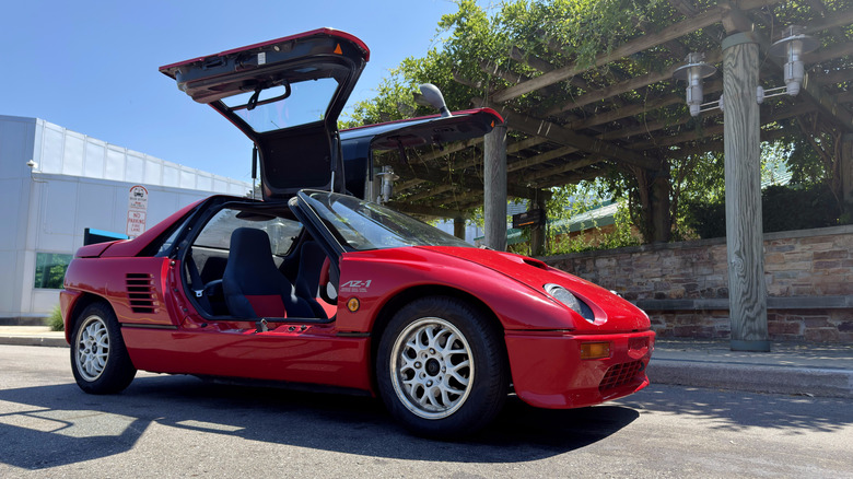Ein roter Mazda Autozam AZ-1 mit geöffneten Flügeltüren parkte vor einem Gebäude mit einem grün beladenen Gitter davor.