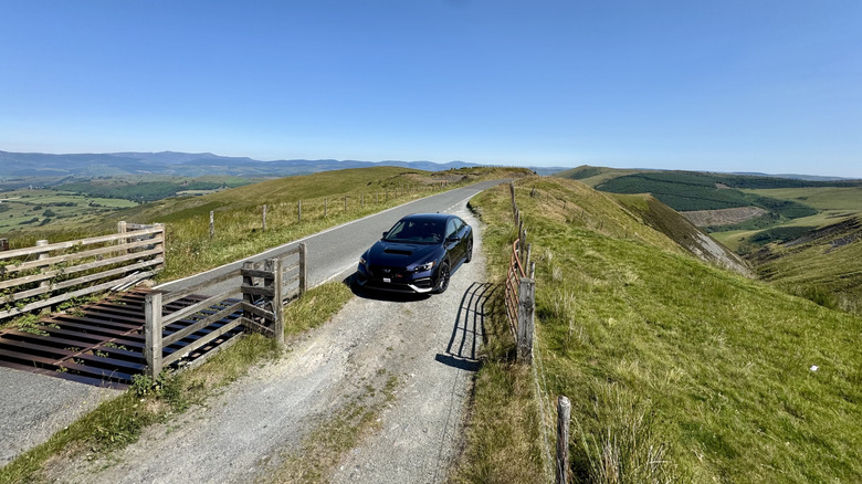 Eine Draufsicht eines Galaxy Purple Pearl WRX, der auf einer Kiesweiche neben einem Kuhfänger auf dem Gipfel eines grasbewachsenen Berges geparkt ist, mit Blick auf Wiesen und blauen Himmel