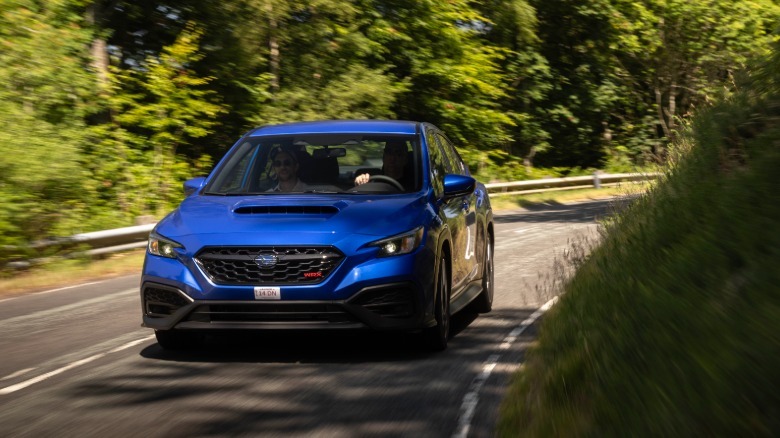 Eine Frontalaufnahme eines blauen WRX, der auf einer von Bäumen und Gras umgebenen Landstraße um eine Ecke fährt