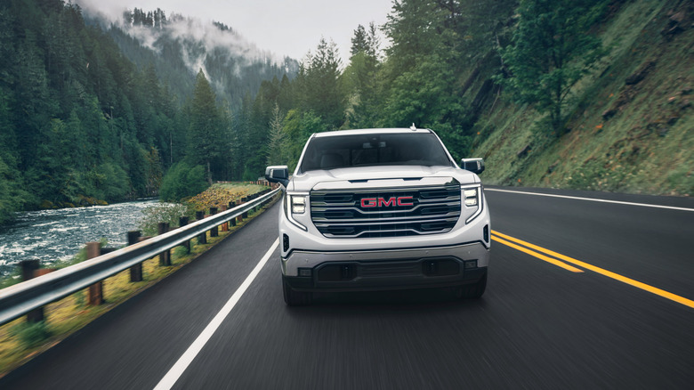Vorderansicht eines weißen 2024 GMC Sierra 1500, der durch einen bergigen Wald neben einem Fluss fährt