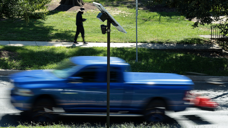 Eine Axon ALPR-Kamera und ein Solarpanel sind an einem Mast neben einer Straße montiert, während ein blauer Pickup und ein Fußgänger auf einem Bürgersteig vorbeifahren