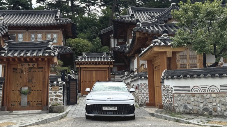 Eine Frontalaufnahme des weißen Grandeur, der zwischen zwei alten traditionellen koreanischen Häusern vor riesigen Kiefern geparkt ist