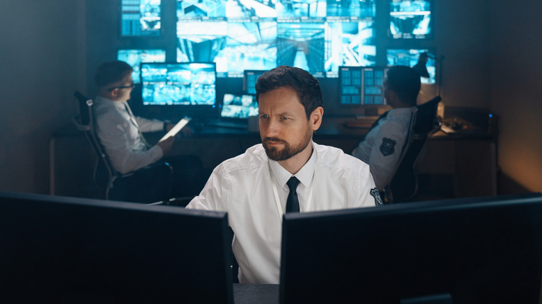 Ein uniformierter Mann sitzt vor Computerbildschirmen mit anderen Beamten und großen Bildschirmen im Hintergrund