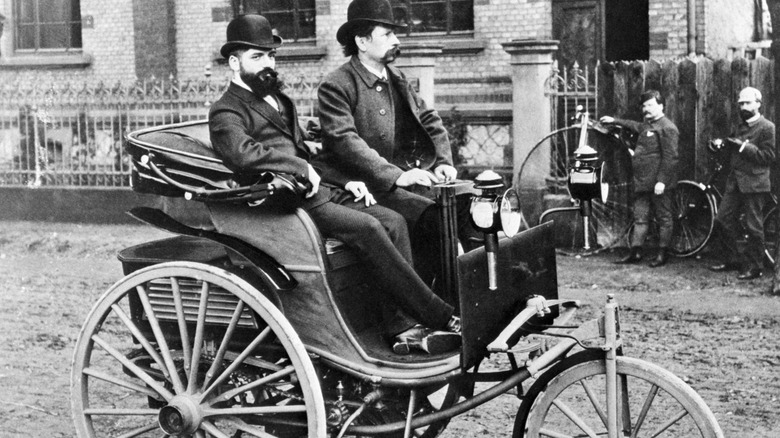 Ein Schwarz-Weiß-Bild von zwei Personen im ersten Mercedes-Benz, einem Einzylinder-Dreirad, mit zwei weiteren Personen im Hintergrund in der Nähe von Fahrrädern.