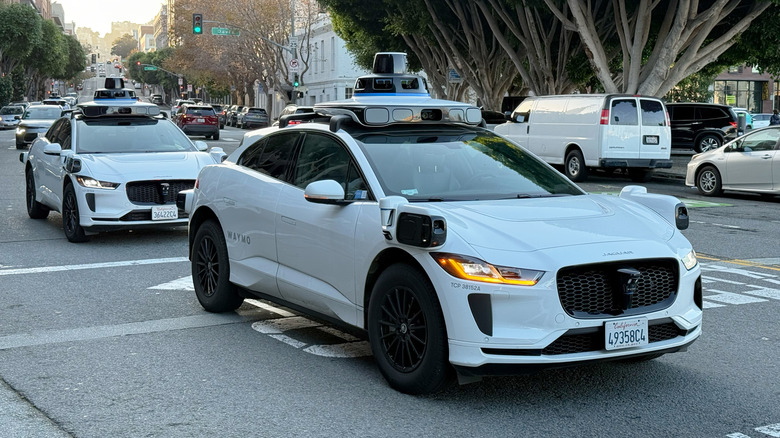 Vordere 3/4-Ansicht von zwei Waymo-Robotaxis, die an einer Kreuzung in der Stadt nach rechts abbiegen