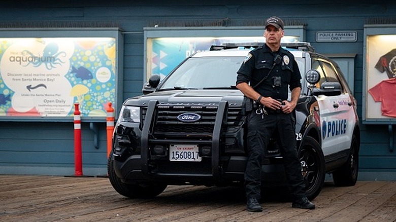 Ein Polizist mit ernstem Gesichtsausdruck neben seinem Ford Police Interceptor SUV, der den Santa Monica Pier im Auge behält.