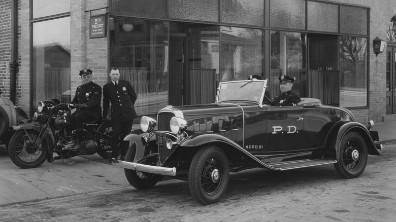 Ein Schwarzweißfoto von Beamten in einem Polizeiauto und auf einem Motorrad vor einer Polizeistation im Jahr 1929.