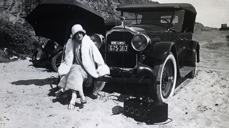 Eine Frau sitzt auf der vorderen Stoßstange eines geparkten alten Autos, neben ihr liegt ein großes altes Radio auf dem Boden