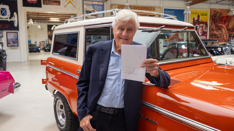 Jay Leno steht mit dem Ellbogen auf dem orangefarbenen Bronco in seiner Garage und hält die unterschriebene Rechnung
