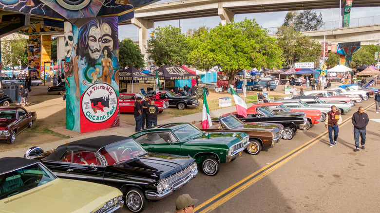 San Diego, Kalifornien – 25. April 2026: Eine Drohnenaufnahme aus geringer Höhe restaurierter Oldtimer und Lowrider mit zur Ausstellung geöffneten Motorhauben. Die Teilnehmer versammeln sich unter der Coronado Bridge zur 56. jährlichen Feier zum Chicano Park Day im Barrio Logan, San Diego. von: Oak City Drone