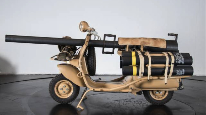Eine sandfarbene Vespa 150 TAP, ausgestattet mit einer Waffe, in einem Studio mit weißer Wand und schwarzem rundem Tisch.