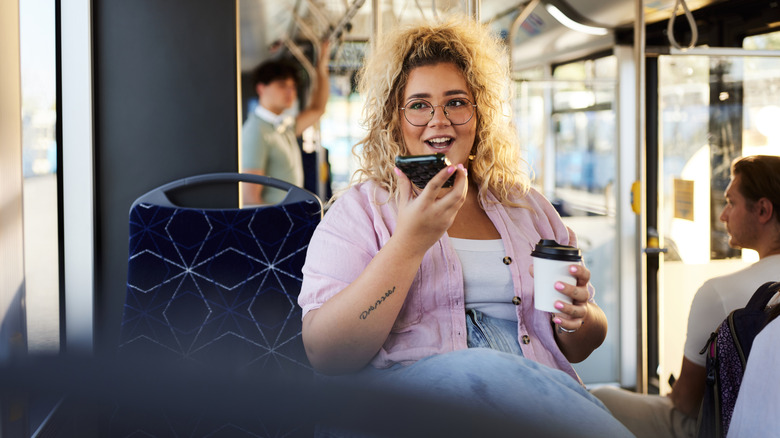 Eine größere blonde Frau mit Brille telefoniert über den Lautsprecher, während sie in einem öffentlichen Bus fährt.