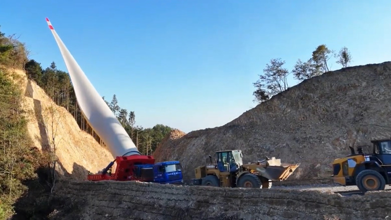 Zwei gelbe Raupenbagger weisen einem kleinen Windturbinenschlepper den Weg, dessen Schild um mindestens 60 Grad angehoben ist, während er auf einem schmalen Pass neben einer Sanddüne eine Kurve fährt.