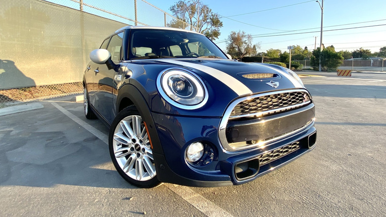 Vordere Dreiviertelaufnahme meines blauen Mini mit weißen Streifen auf der Motorhaube, geparkt auf Asphalt vor blauem Himmel und grünem Zaun
