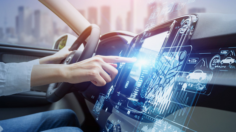 Ein Finger zeigt auf den Infotainment-Bildschirm eines Autos, der von projizierten Datenillustrationen umgeben ist.