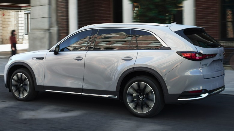 Hintere Dreiviertelaufnahme eines silbernen CX-90, der an einem Backsteingebäude vorbeifährt